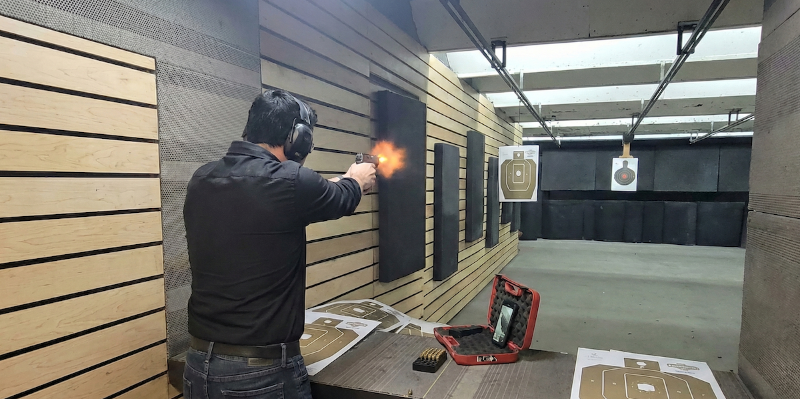 Sie sehen einen Pistolenschützen auf einem Indoor-Schießstand beim Schuss, mit Mündungsfeuer und Zielscheiben im Hintergrund.
